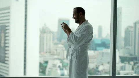 Young man in bathrobe playing game on smartphone standing by window at home, 4K Stock Footage 94860351