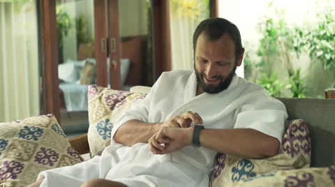 Young man in bathrobe sitting with smartwatch on sofa in outdoor villa Видео 63345175