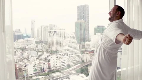 Young man in bathrobe stretching his arms by window with city view, super slow m Stock Footage 127974019