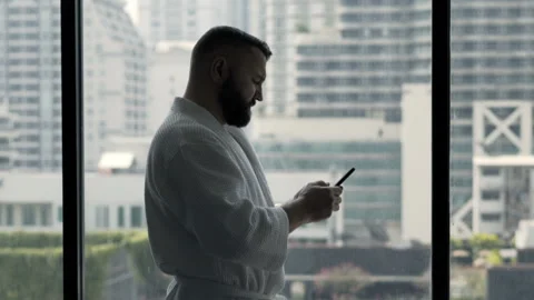 Young man in bathrobe texting on smartphone standing by window at home Stock Footage 159065521