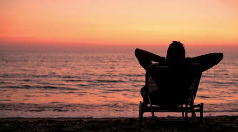 A young man on the beach lying in lawn chair watching the sunset Stock Footage 33847617
