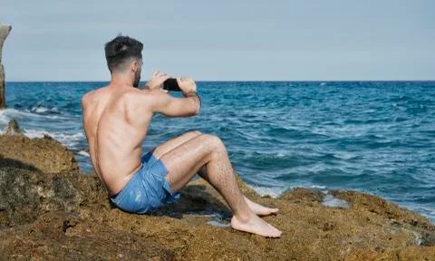 Young man on beach using cell phone to film the sea Stock Photos