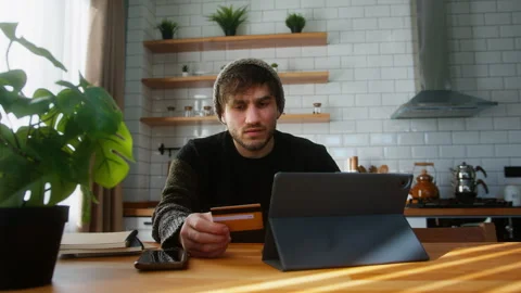 Young man with beanie sitting in kitchen at home entering credit card numbe.. Stock Footage 261164900