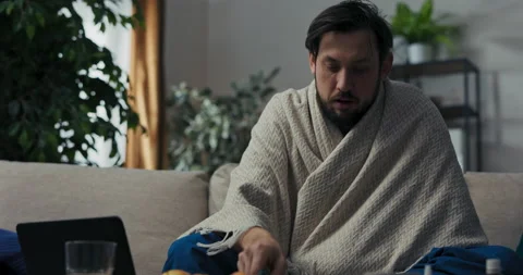 Young man with beard and black hair is sitting on sofa, taking temperature and 스톡 동영상 218329908