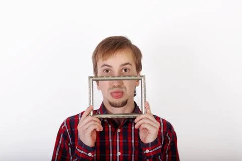 Young man with beard in checkered red shirt shows tongue behind frame Stock Photos