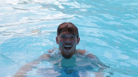 A young man with a beard dives and comes up with a splash in the pool with clear Stock Footage 70774203
