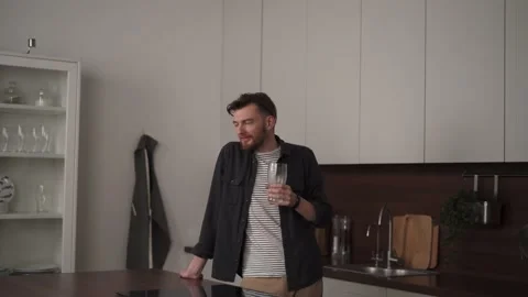 The young man with the beard drinks a glass of water in his kitchen Stock Footage 169046494