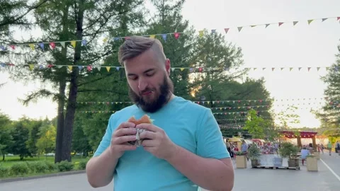 Young man with beard eats and chews burger at summer streetfood festival Stock Footage 229196256
