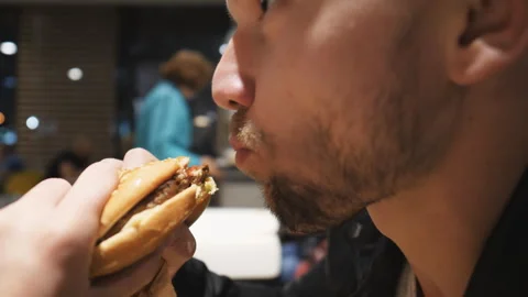 Young man with beard eats hamburger or cheeseburger at food court in shopping 库存影片 333693016