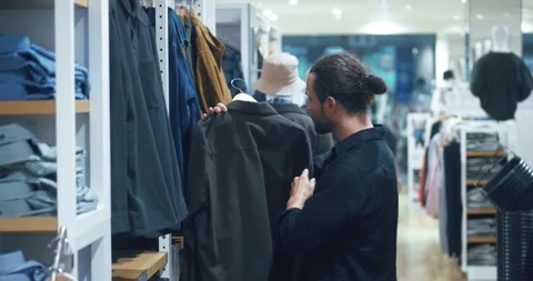 Young man with beard examines stylish jackets, people choose clothes in order to Stock Footage 257701991