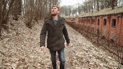 A young man With a beard, in a gray coat walks through the park in the spring or Stock Footage 74136254