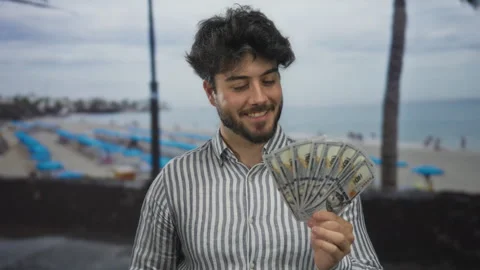 Young man with a beard holding us dollars, smiling at the seaside, with umb.. Stock Footage 310993142