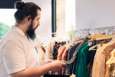 Young man with beard, looking at the labels of clothes offers, in fashion shop Stock Photos
