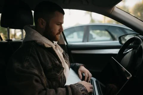A young man with a beard opens his laptop computer to do business inside a ca Stock Photos