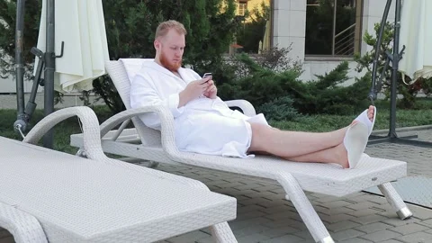 Young man with beard sits by hotel pool on vacation with a phone in his hands Video stock 147038564