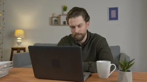 Young man with a beard sits at a table, works on a laptop, works at home, smiles Stock-Footage 128016734