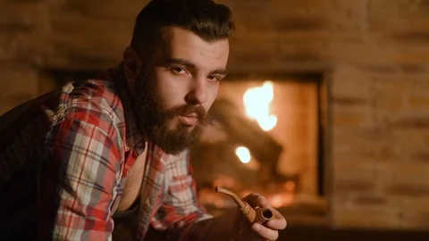 A young man with a beard smokes a pipe in a wooden house 動画素材 72465456