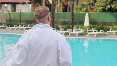 Young man with beard strips off, takes off white robe by the hotel pool Stock Footage 147041158