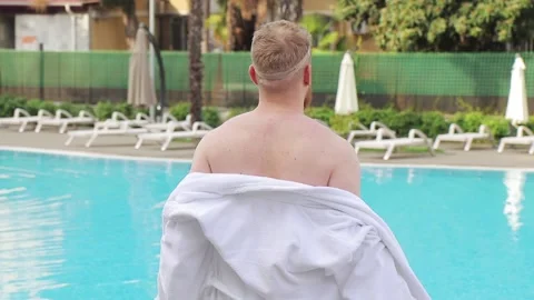 Young man with beard strips off, takes off white robe by the hotel pool Stock Footage 147041443