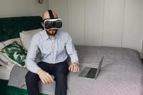 Young man with a beard in virtual reality glasses sits on the edge of the bed Stockfoto's