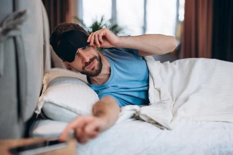 Young man with beard waking up in bed reaches out to turn off alarm on mobile Stock Photos