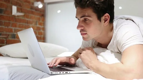 Young Man in Bed Working on Laptop and Reacting to Success Stock Footage 83805020