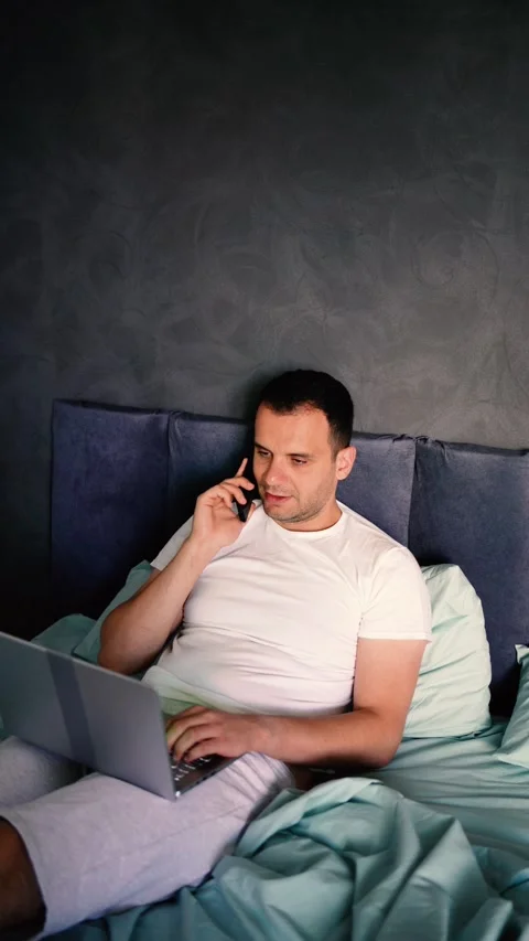 Young man on the bed working on a laptop and talking on the phone Stock Footage 276379501