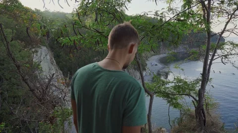 A young man from behind goes to the cliff and looks at the bottom. Stockbeeldmateriaal 117628886