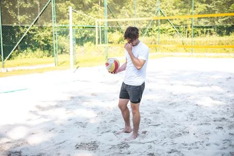 Young man bends down to pick up a volleyball from the sandy court, preparing  Stock Photos