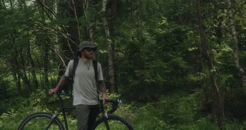 Young man with bicycle standing on forest road, looking around Stock Footage 311934917