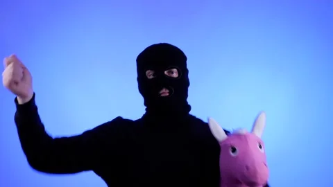 Young man in black balaclava dancing and fooling around with pink toy horse Stock Footage 130596433