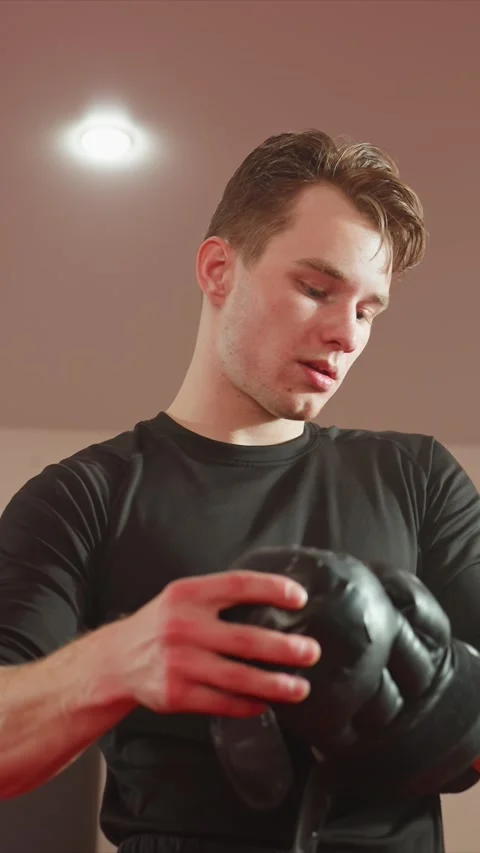 Young man in black boxing attire adjusting gloves preparing for exercise indoors Stock Footage 343092482