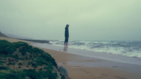 Young man in black clothes stands on the ocean shore, waves. Man stands in fr Stock Footage 197336715