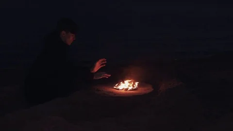 Young man In black jacket seating near bonfire of burning shoes on sand at coast Stock Footage 74380392