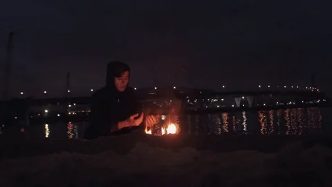 Young man In black jacket seating near bonfire of burning shoes on sand at coast Stock Footage 74380531