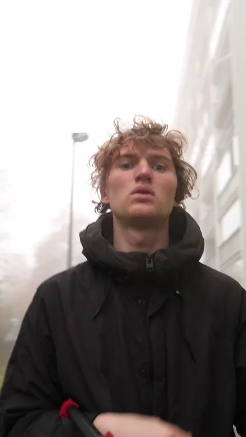 A young man in a black jacket stands in front of a building, looking around as i Stock Footage 290187187