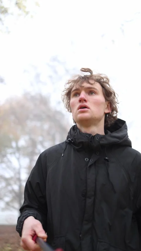 A young man in a black jacket stands next to a tree, looking off into the distan Видео 290187403