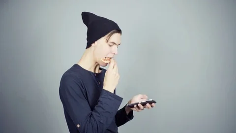Young man in black knitted hat eating pizza using smartphone gray background Stock Footage 70260984