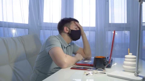 A young man in a black mask sits on a white sofa at a laptop Vidéo 130001246