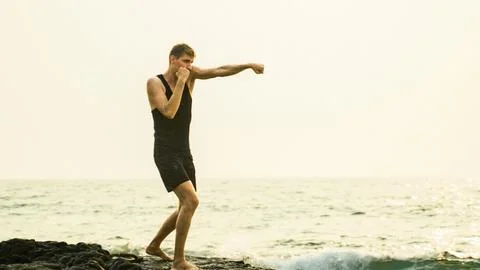 Young man in a black sports suit is boxing against the backdrop of the sea Stock Photos