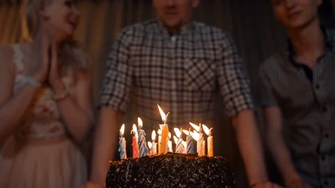 Young man blows out candles on a festive cake with friends near. 96528060