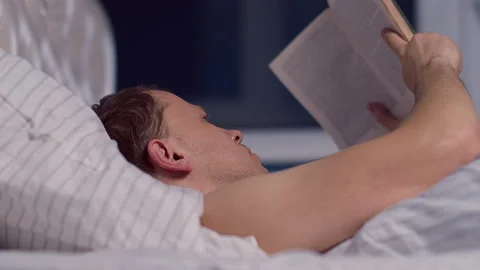 A young man with a book lying in bed at night before going to bed Stock Footage 132722937