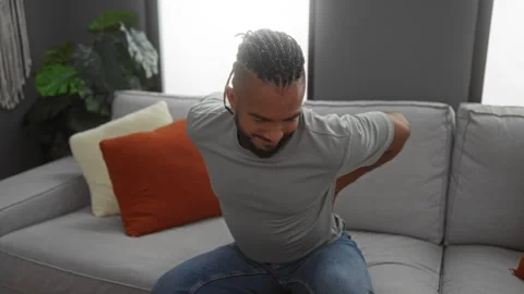 Young man with braids experiencing back pain while sitting on a comfortable.. Vidéo 301691415