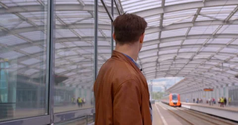 A young man in a brown leather jacket stands on the platform of the ground metro Stock Footage 119632437