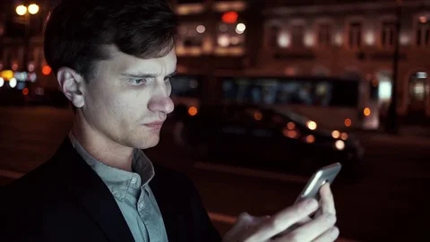 Young man browsing his cellphone on a busy urban street at night Stock Footage 80009764