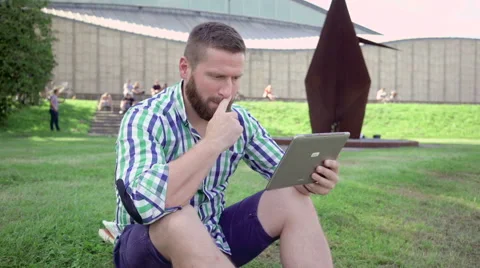 Young man browsing tablet, sitting on grass. Steadicam. Vídeo Stock 67236311