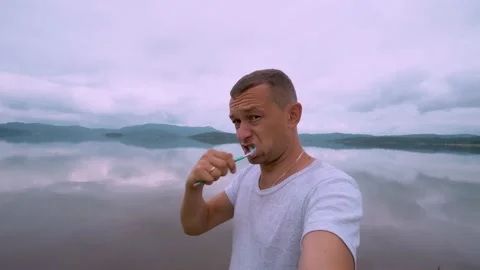 Young man brushes his teeth while standing outdoors on hill with lake mountains. Stock Footage 204992153