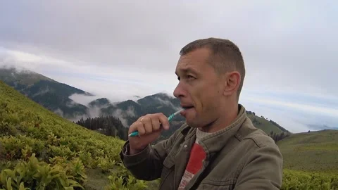 Young man brushes his teeth while standing outdoors on hill with mountains. Stock Footage 204993432
