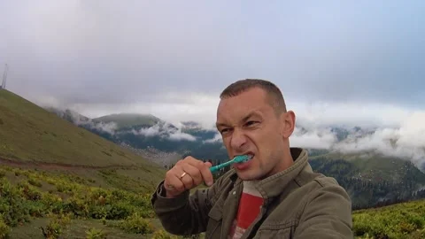 Young man brushes his teeth while standing outdoors on hill with mountains. Stock Footage 204993567