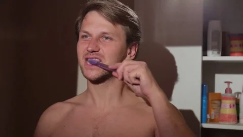 Young man brushing his teeth. He is standing topless in the bathroom. Stock Footage 76116354